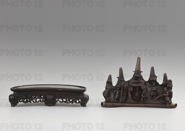 Brush stand on base;, Chinese; Qing dynasty 1644–1911; 19th century; painting implements; subject: mountains, men, horses, scholars; depiction: Figure of a Hermit in a cave and other figures; materials/technique: varia; dimensions: height 21.0 cm, width 10.0 cm, depth 25.0 cm., A low, dark brown wooden stand with an elongated oval top and ornate openwork apron sits to the left, showing carved scrolling patterns and short curved feet; to the right is a dark brown carved wooden sculpture resembling a jagged mountain ridge with several peaked forms and carved human figures integrated into its surface, including standing figures and a seated figure within a recessed area, all on a flat base; the background is a smooth gradient of gray fading to a lighter gray near the bottom, and the surface beneath the objects is a pale off-white.