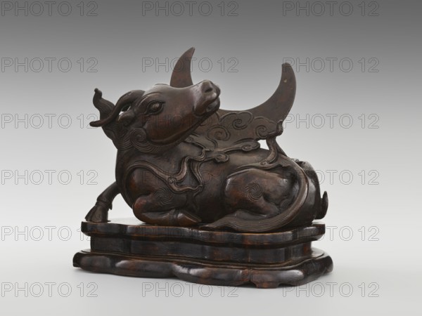 Mirror pedestal - lying unicorn on base; maker, Chinese; Qing dynasty 1644–1911, 17th century; carved wooden base; subject/iconography: qilin unicorn, crescent; function: mirror pedestal / toilet accessory; dimensions: height 16 cm, width 11.5 cm, depth 19.5 cm., A carved sculpture of an animal reclining on a layered base, rendered in a dark brown wood with visible lighter brown highlights and some blackened areas; the animal has a rounded body with stylized flowing mane and tail details, curled decorative motifs on its flanks and back, a raised head with pronounced eyes and ears, and a curved crescent-shaped element rising behind it; the base is shaped with rounded edges and shows similar dark brown and black tones with worn lighter brown patches, and the background is a smooth gradient of gray.