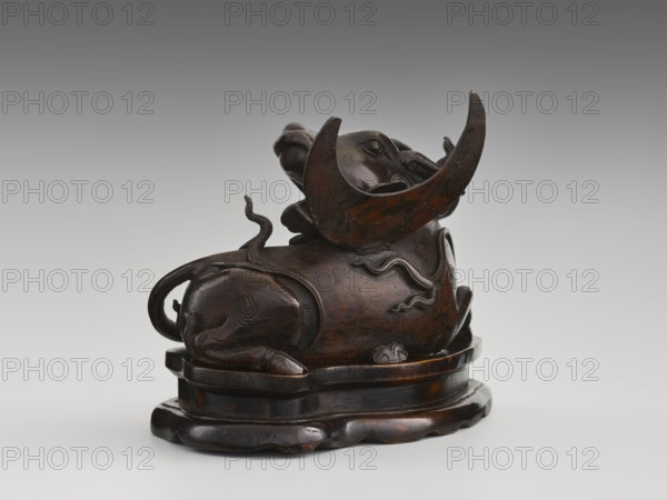Mirror pedestal — lying unicorn on base; Chinese; Qing dynasty 1644–1911, 17th century; carved wooden base; subject/iconography: qilin unicorn, crescent symbol; function: mirror pedestal / toilet accessory; materials: carved wood; dimensions: height 16 cm, width 11.5 cm, depth 19.5 cm., A dark brown carved object sits on a rounded base, showing a sculpted reclining animal form with a curled tail and raised head. The surface has a polished, glossy finish with lighter brown highlights and subtle black areas, and there are flowing carved lines and raised decorative motifs along the body. A large crescent-shaped element arches near the head, and a few sinuous, ribbon-like forms rise from the back. The base and the carved piece share similar dark brown and black tones with occasional warmer brown patches.