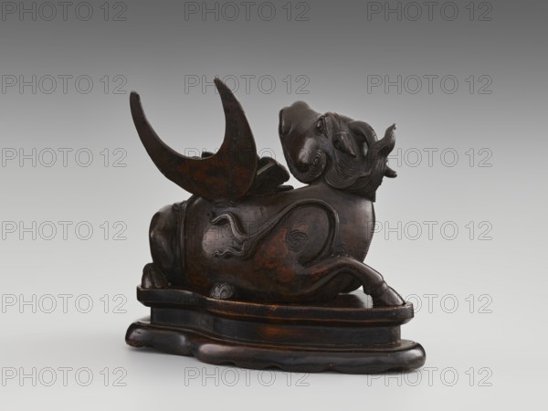 Mirror pedestal — lying unicorn on base; carved wooden base; qilin unicorn iconography; crescent symbol; mirror; toilet accessory; Chinese art; Qing dynasty 1644–1911; 17th century; China; height 16 cm, width 11.5 cm, depth 19.5 cm., A dark brown carved object resembling a reclining mythical animal with a textured mane and curled tail sits on a carved base; the creature has a rounded body with flowing decorative reliefs, a raised head with stylized facial features, and a large curved crescent-shaped element attached near its back. The piece shows varying brown tones with areas of darker nearly black patina and subtle reddish-brown highlights, placed against a smooth gradient gray background.