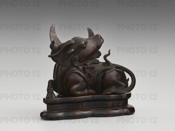 Mirror pedestal — lying unicorn on base; Chinese; qilin iconography; unicorns iconography; carved wooden base; mirrors; crescent symbol; Qing dynasty 1644–1911; 17th century; China; toilet accessory; height 16 cm, width 11.5 cm, depth 19.5 cm., A dark brown carved sculpture of a reclining animal-like figure with curved horns and stylized flowing mane details, shown on a rounded rectangular base; the figure has smooth, rounded body contours, a curled tail with textured lines, raised ears, and decorative carved swirls on its shoulders and hindquarters. The colors visible are dark brown with areas of lighter brown highlights and a neutral gray gradient background.