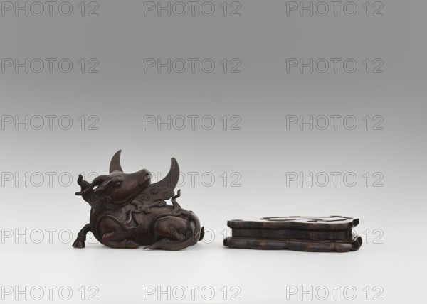 Mirror pedestal — lying unicorn on base; Chinese; Qing 1644–1911, 17th century; carved wooden base; subject: qilin/unicorn iconography and crescent symbol; function: mirror pedestal / toilet accessory; materials: carved wood; dimensions: height 16 cm, width 11.5 cm, depth 19.5 cm., A dark brown carved object resembling a reclining animal with stylized horns and flowing decorative motifs lies on a light surface, showing smooth rounded contours, carved details, and a polished finish; nearby is a separate dark brown carved base with layered, organic edges and a similar polished appearance, all set against a gradient gray background.