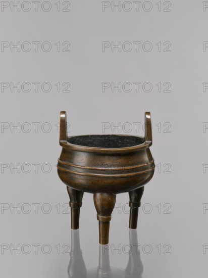 Ding-type incense burner on a stand, Qing dynasty 1644–1911, 19th century, China. Bronze ceremonial vessel decorated with lotus motifs, stylized FU happiness hieroglyphs, lotuses, bats, fo, tiger, meander ornament, and the one hundred antiquities baigu motif. Round wooden turned stand with five legs on a pedestal shaped like a lotus rosette. Function: ritual/ceremonial incense burner. Dimensions: incense burner height 21 cm, diameter 15 cm; base height 7.5 cm, width 18.4 cm., A bronze tripod vessel with a round body and a dark interior sits against a pale gray background; the vessel has a warm brown and dark bronze patina with subtle etched patterns on its surface, a flared rim supported by two short upright handles, and a ringed band encircling the upper and midsection, standing on three tapered legs that reflect faintly on the glossy surface below.