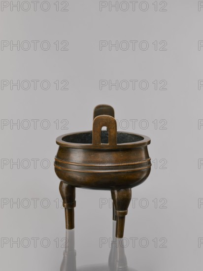 Ding-type incense burner on a stand;, 19th century, China, Qing dynasty 1644–1911. Bronze ceremonial ritual vessel incense burner decorated with lotus motifs, bats iconography of happiness, fo, tiger symbol, meander and one hundred antiquities/baigu ornament, stylized hieroglyphs reading FU happiness. Round turned wooden stand with five legs on a pedestal shaped as a lotus rosette. Incense burner: height 21 cm, diameter 15 cm. Stand base: height 7.5 cm, width 18.4 cm., A bronze tripod vessel with a rounded body and a concave rim sits against a neutral gray background, showing a warm brown and dark bronze patina with subtle greenish-black interior tones; the vessel rests on three slightly tapered legs that reflect on the surface below, and a vertical handle with a rounded top and an arched cutout rises from the rim, all surfaces displaying faint engraved patterns and a smooth, aged finish.
