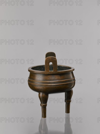 Ding-type incense burner on a stand, Bronze ceremonial ritual vessel, Qing dynasty 1644–1911, 19th century, China. Decoration: lotus motifs; bats iconography; fo; tiger symbol; meander and one hundred antiquities baigu ornament; stylized hieroglyphs FU happiness. Round wooden turned stand with five legs, on a pedestal shaped like a lotus rosette. Measurements: incense burner height 21 cm, diameter 15 cm; base height 7.5 cm, width 18.4 cm., A bronze tripod vessel with a rounded body and a flared rim stands on three tapered legs; the surface shows a dark brown to bronze patina with areas of darker brown and subtle greenish-black interior shading, and the body is decorated with incised patterns; a tall arch-shaped handle rises from the rim and shares the same bronze and brown tones; the background is a smooth light gray.