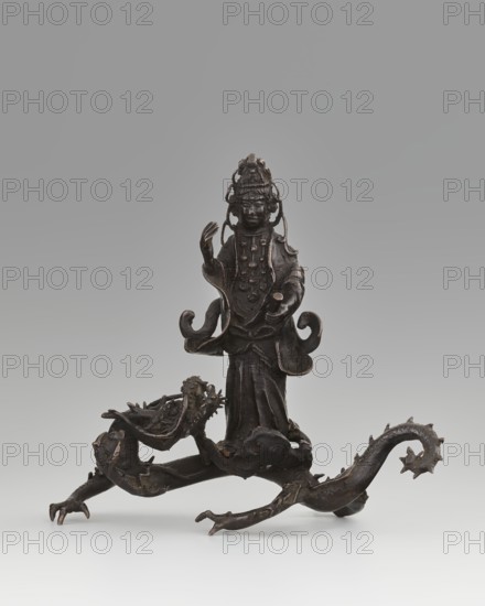 Guanyin on a dragon;, Chinese; Qing dynasty 1644–1911, 18th century; sculpture, figurine; materials not specified; subject: Guanyin bodhisattva of mercy riding a dragon with a pearl; iconography: dragons, Buddhism, goddesses; dimensions: overall height 15 cm, width 7.1 cm; dragon element height 4 cm, width 8.3 cm., A dark bronze figurine stands on a sculpted base formed by a sinuous dragon-like creature, with a few curling limbs and clawed feet extending outward. The standing figure wears layered garments with flowing sleeves and rounded ornamental details on the chest, and a tall, elaborate headdress with looping elements that frame the face. One arm is raised with an open hand while the other extends forward holding a small cylindrical object. The dragon-like base has a textured head with an open mouth and ridged spine, and a curled tail with pointed projections. The visible colors are dark brown and black tones with subtle lighter highlights where the metal catches light, set agains...