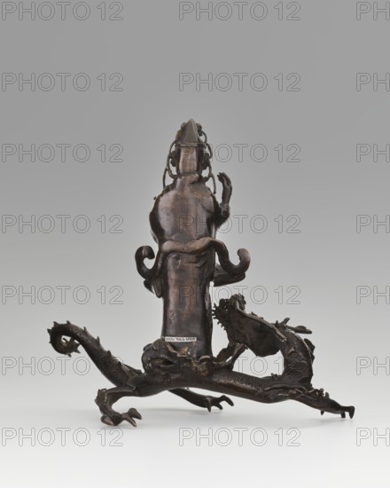 Guanyin on a dragon;, gift; Chinese art; Chinese; Qing 1644–1911; dragons iconography; Buddhism; bodhisattva iconography; goddesses; mercy; dragon with a pearl; 18th century; China; sculpture, figurine; overall height 15 cm, width 7.1 cm; dragon: height 4 cm, width 8.3 cm., A dark brown carved object with a glossy patina sits against a light gray background; it stands upright on a base formed by a sculpted creature with a sinuous body, curled tail, clawed feet, and a textured surface suggesting scales, while the upright portion rises from the base with flowing, rounded forms and curled decorative elements on its sides; the object shows varying brown tones from deep near-black to lighter warm brown highlights on raised areas, and the scene includes subtle shadows beneath the piece on a pale surface.