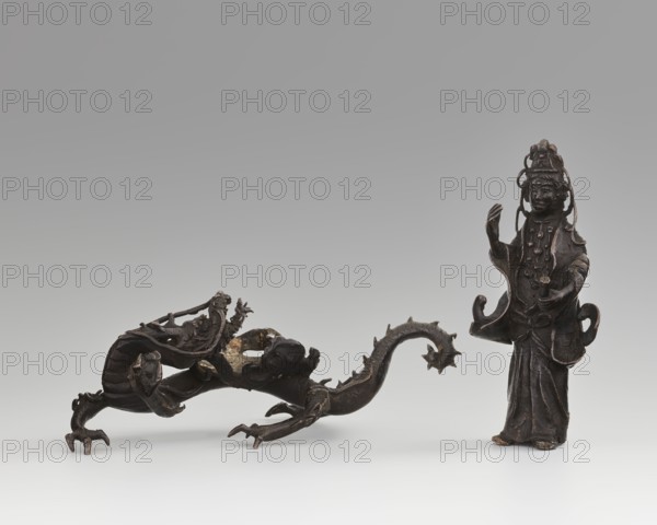 Guanyin on a dragon;, ; Chinese art; Chinese culture; Qing dynasty 1644–1911; dragons iconography; Buddhism; bodhisattva iconography; goddesses; mercy; dragon with a pearl; 18th century; China; sculpture, figurine; dimensions: overall height 15 cm, width 7.1 cm; dragon: height 4 cm, width 8.3 cm., A dark bronze sculpted figure of a robed person stands with one hand raised and the other holding a short cylindrical object, wearing a tall ornate headdress and layered garments with flowing sleeves and decorative elements; beside the figure is a long sinuous dark bronze dragon-like creature with a curved spiked tail, clawed feet, an elongated body and head with open mouth and detailed textured scales, both objects resting on a neutral light gray surface against a smooth gradient gray background, with visible colors including dark brown, black, gray and subtle bronze highlights.