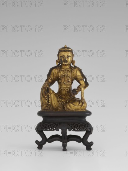 Bodhisattva Manjushri, on a base. Chinese; Qing dynasty 1644–1911. Wood sculpture with wooden stand in the form of a table. 18th century. Bodhisattva holds a rosary in his right hand and a sutra in his left. Sculpture. Height 14.5 cm, width 8.1 cm, depth 10.3 cm. Chinese Buddhism; Chinese/Southeast Asian art., A gilded seated figure with detailed ornamentation and draped garments sits on a carved dark wooden stand; the figure's gold surface shows sculpted jewelry, a headdress, and folded limbs with one hand resting on a knee and the other holding a small rounded object, and the stand is dark brown with ornate openwork and curved legs against a neutral gray background with tones of light gray and white.