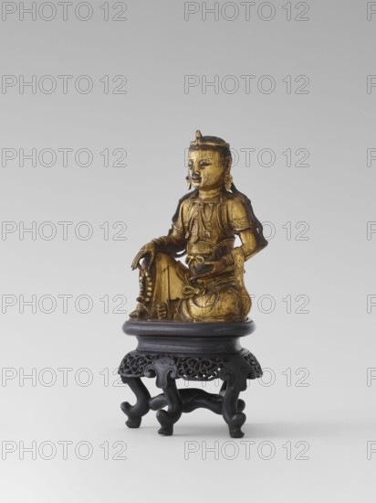 Bodhisattva Manjushri, on a base. Chinese art; Chinese/Southeast Asian, Qing dynasty 1644–1911. Wooden sculpture; Bodhisattva holds a rosary in his right hand and a sutra in his left. Wooden stand in the form of a table. 18th century. Sculpture. Height 14.5 cm, width 8.1 cm, depth 10.3 cm., A gilded figurine of a seated figure with a serene facial expression sits on a dark carved wooden stand; the figure wears ornate garments and jewelry with detailed patterns, has earrings and a headpiece, and holds a string of beads in one hand while the other hand rests near a small bowl; the gilded surface shows areas of darker patina, and the stand is a deep brown or black with carved openwork and curved legs; the background is a smooth light gray.