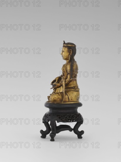 Bodhisattva Manjushri, on a base. Chinese art; Chinese/Southeast Asian, Qing 1644–1911. Figurine of a Bodhisattva holding a rosary in his right hand and a sutra in his left. Wooden stand in the form of a table. Sculpture, 18th century. Height 14.5 cm; width 8.1 cm; depth 10.3 cm., A small gilded figure seated on a carved dark wooden stand, shown in profile facing right, with long hair pulled back and a simple headband, wearing layered garments and holding an object in both hands; the figure's surface shows areas of worn gold and darker underlying tones, and the stand has a round top, ornate pierced apron, and curving legs in a dark brown or black color, all set against a pale gray background.