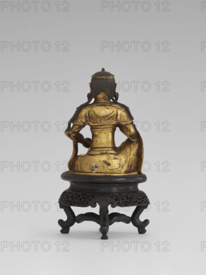 Bodhisattva Manjushri, on a base. Chinese art; Chinese/Southeast Asian culture. Qing dynasty 1644–1911. Wood sculpture/figurine, 18th century. Bodhisattva holds a rosary in his right hand and a sutra in his left. Wooden stand in the form of a table. Height 14.5 cm; width 8.1 cm; depth 10.3 cm., A gilded bronze seated figure shown from the back, with a dark patina on the hair and edges, wearing sculpted garments with decorative raised patterns, seated on a rounded dark wooden pedestal that has carved openwork around its rim and curved legs; visible colors include gold, dark brown, and black against a neutral gray background.