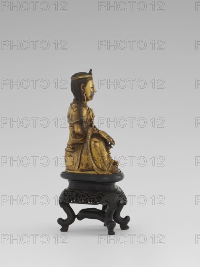 Bodhisattva Manjushri, on a base. Chinese, Qing dynasty 1644–1911, 18th century. Wooden sculpture; Bodhisattva holds a rosary in his right hand and a sutra in his left. Wooden stand in the form of a table. Height 14.5 cm; width 8.1 cm; depth 10.3 cm. Cultural context: Chinese/Southeast Asian, Chinese Buddhism., The image shows a small gilded figurine of a seated person with long dark hair and a headband, rendered in gold and darker patinated tones, positioned on a carved dark wooden stand with an ornate openwork apron and curved legs; the background is a smooth gradient of light gray to slightly darker gray.