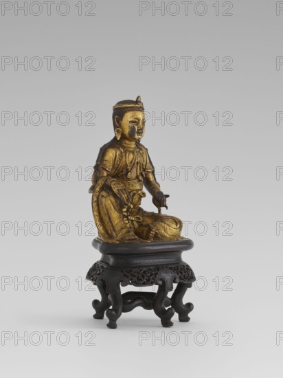 Bodhisattva Manjushri, on a base. Chinese Buddhism; Chinese art; Asian art. Qing dynasty 1644–1911. Wooden sculpture with a wooden stand in the form of a table. Bodhisattva holds a rosary in his right hand and a sutra in his left. 18th century. Height 14.5 cm; width 8.1 cm; depth 10.3 cm. Culture: Chinese/Southeast Asia place of origin uncertain., A gilded figurine of a seated figure with detailed ornamentation and a calm facial expression rests on a dark carved wooden stand; the figure wears a headdress and earrings, draped garments with intricate patterns, and has one hand extended while the other rests near the lap, showing sculpted folds and decorative elements in gold tones contrasted with the black-brown of the stand and a pale gray background.