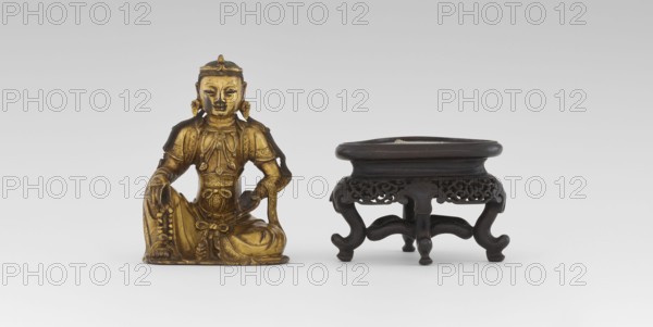 Bodhisattva Manjushri, on a base. Chinese art; Chinese, Southeast Asian; Qing dynasty 1644–1911. Figurine of Chinese Buddhism: the Bodhisattva holds a rosary in his right hand and a sutra in his left. Wooden stand in the form of a table. 18th century; sculpture. Height 14.5 cm, width 8.1 cm, depth 10.3 cm., A small gilded statue of a seated figure with ornate clothing and jewelry sits next to a dark wooden low table or stand with carved openwork sides and cabriole legs; the statue shows detailed patterns and drapery and a serene facial expression, and the stand has a slightly rounded top and curved supports. Colors visible include gold on the statue and dark brown to nearly black on the wooden stand, set against a pale gray to white background.