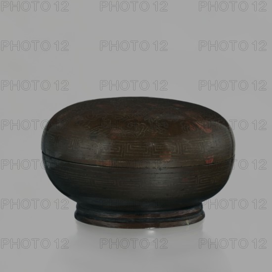 Box with a lid; Chinese; Qing dynasty 1644–1911, 18th century; vessel for writing implements; decoration of dragons dragon with a pearl, inlaid with silver thread; height 4.5 cm, diameter 7.2 cm., A rounded, low-profile container with a lid sits centered against a light gray background, showing a dark brown surface with patches of reddish-brown and subtle darker markings; the lid and base display faint linear and geometric carved patterns, and the piece rests on a short circular foot with slightly lighter brown wear around its edge.