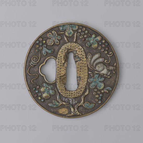 Crossguard tsuba, Japanese, Edo period 1603–1868, 17th century. Round tsuba maru-gata with slightly raised uchi-kaeshi type edge and oval seppa-dai. Dark patinated background highlighting golden brass on the edge, around kôgai hitsuana and seppa-dai. Relief decoration in colored enamels of a grape vine and a squirrel on both sides. Unsigned. Japan; diameter 6.9 cm, thickness 0.5 cm., A round metal object with a central elongated keyhole and a decorative irregular cutout, featuring raised floral and vine motifs and a small bird-like figure; the surface shows areas of gold-toned metal, dark brown or black patina, and turquoise-green and pale blue enamel accents, with touches of reddish and yellowish hues in some floral elements.