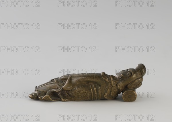Paperweight: figurine of a lying/sleeping woman on a pillow, Chinese, Qing dynasty 1644–1911, 19th century; materials/attributes include painting implements; dimensions height 2.5 cm, width 2.7 cm., A small reclining figure sculpture resting on its side on a pale background, rendered in a metallic bronze color with darker brown and greenish patina areas; the figure has carved ornamental details on its clothing and a rounded head with stylized hair or headdress, and it leans against a cylindrical support piece that shares the same bronze tones.