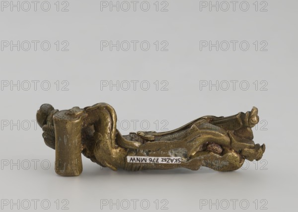 Paperweight - lying woman; Chinese, Qing dynasty 1644–1911, 19th century; figurine of a sleeping woman on a pillow; materials: painting implements, paperweight; dimensions: height 2.5 cm, width 2.7 cm., A small sculptural object lies on a neutral gray background, appearing metallic with a worn surface and a patina of gold and brown tones. The object has an irregular, organic shape with flowing, curved forms and textured details, showing areas of darker brown and greenish discoloration. One end features a cylindrical protrusion and a rounded knob-like element, while the body includes layered, undulating ridges and recessed cavities. There are also small patches of darker rust-colored spots and some areas where the metallic finish looks scuffed or abraded.
