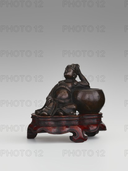 Paperweight — Li Bai on a stand; sculptor, ; Chinese art; Chinese culture; Li Bai 701–762; figurine, paperweight; Qing dynasty 1644–1911; alcohol vessel; poet; painting implements; 19th century, China; sculpture; height 6.3 cm, width 8.0 cm, A small dark brown carved figure of a seated person leaning against a rounded vessel sits on an ornately shaped low wooden stand with a glossy reddish-brown finish; the figure appears relaxed with one arm resting on the vessel and the other hand touching the head, showing folds in the clothing and rounded facial features, and the composition contrasts the deep brown of the figure and vessel with the warmer red tones of the stand against a neutral gray-to-white background.