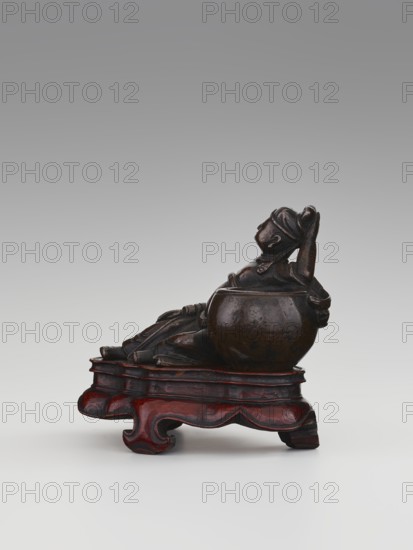 Paperweight depicting Li Bai on a stand; sculptor unknown. Chinese, Qing dynasty 1644–1911, 19th century. Materials/objects: sculpture/figurine paperweight; related to alcohol vessels, poets, and painting implements. Dimensions: height 6.3 cm, width 8.0 cm., A dark brown carved figure reclining against a rounded vessel sits on an ornately shaped wooden stand that is reddish-brown with darker accents; the figure appears to wear a headwrap and loose clothing and has a glossy surface with lighter brown highlights, while the background is a smooth gradient of gray transitioning from darker at the top to lighter toward the bottom.