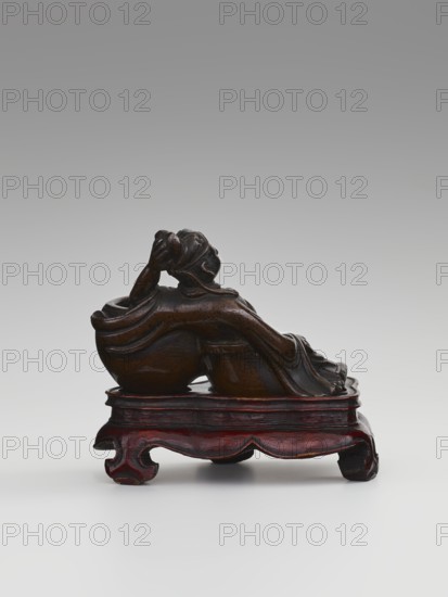 Paperweight — Li Bai on a stand; sculptor unknown. Chinese, Qing dynasty 1644–1911, 19th century. Figurine/stone sculpture used as a paperweight; depicts the poet Li Bai 701–762 on a stand. Materials: sculpture likely stone; function: paperweight; related forms: alcohol vessels, painting implements. Dimensions: height 6.3 cm, width 8.0 cm., A small dark brown carved figure reclining on a carved wooden stand, the figure showing flowing drapery and a detailed head turned upward with a hand raised to the face, resting on a reddish-brown stand with curved feet and a slightly glossy finish, set against a neutral gray to white gradient background.