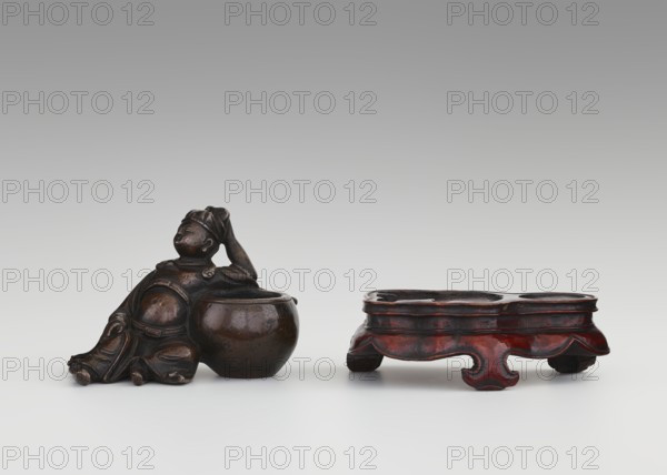 Paperweight: Li Bai on a stand; sculptor unknown. Material: sculpture; subject: Li Bai 701–762 depicted with painting implements and alcohol vessels. Style/period: Qing dynasty 1644–1911, 19th century. Function: paperweight/figurine. Dimensions: height 6.3 cm, width 8.0 cm., A dark brown sculptural figure reclining beside a round, bowl-like vessel sits to the left, the figure showing a rounded torso, draped clothing, and one arm raised to touch the head; to the right is a low, ornate stand with a scalloped, curving edge and short feet, finished in a reddish-brown and dark brown patina; the background is a smooth gradient from light gray at the bottom to darker gray at the top, and the surface beneath the objects is a pale, off-white-gray.