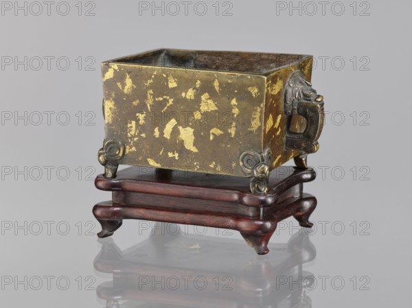 Censer on base, Chinese, Qing dynasty 1644–1911, 18th century. Ceremonial vessel censer with lion/fo iconography and handles; wooden carved quadrangular base. Materials: bronze censer and wood base. Dimensions: censer height 2.2 cm, width 12.3 cm, length 9.7 cm; base height 1.8 cm, width 12.2 cm, length 9.6 cm., A square metal container with a worn gold and dark brown mottled surface sits on a dark reddish-brown wooden stand with curved legs; the metal shows patchy gold areas and darker brown or blackened interior surfaces, and each corner of the container has decorative metallic fittings in a darker bronze or gray tone, including a sculpted handle on one side shaped like a stylized animal head in a darker metal; the wooden stand has a glossy finish and a layered platform with curving edges and short cabriole-style legs, and the whole object is reflected faintly on the smooth light gray surface beneath it against a pale gray background.