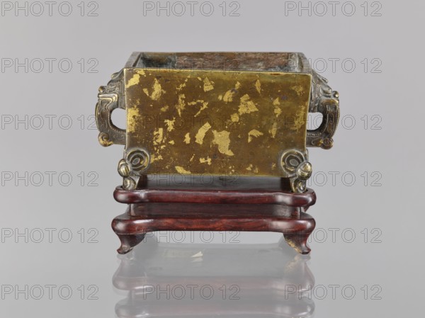 Censer on base;, Chinese, Qing dynasty 1644–1911, 18th century. Ceremonial vessel censer with lions fo iconography and handles. Materials: censer and wooden carved quadrangular base. Dimensions: censer height 2.2 cm, width 12.3 cm, length 9.7 cm; base height 1.8 cm, width 12.2 cm, length 9.6 cm., A rectangular metal vessel with worn gold and brown surfaces sits on a dark reddish-brown wooden stand; the vessel has ornamental handles on each side with a silvery-gray tone and rounded decorative feet at the front, and the interior shows a mix of gray and brown patina. The wooden stand has a polished, curved form with visible grain and darker reddish highlights, and the scene is set against a neutral light gray background with a subtle reflection of the stand and vessel on a glossy surface. The visible colors include gold, brown, dark reddish-brown, silvery-gray, gray, and hints of darker and lighter tones.