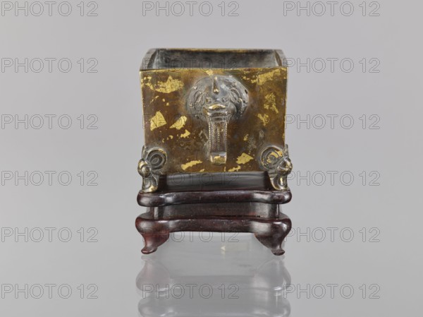Censer on base, Chinese, Qing dynasty 1644–1911, 18th century. Censer vessel with lions fo and handles; ceremonial/ritual vessel in Chinese art. Materials: censer and base; wooden base carved, quadrangular. Dimensions: censer height 2.2 cm, width 12.3 cm, length 9.7 cm; base height 1.8 cm, width 12.2 cm, length 9.6 cm., A square metallic container with a worn gold and brown surface sits on a dark reddish-brown wooden stand with curved legs; the front of the container features a raised decorative element with a rounded face-like shape and a vertical ornament beneath it, and there are small ornamental scroll-like protrusions at the lower front corners; the scene is set against a light gray background and the surface beneath reflects the object.
