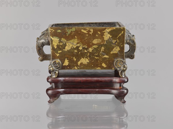 Censer on base, Chinese, Qing dynasty 1644–1911, 18th century. Ceremonial vessel censer with lions fo iconography and handles; wooden carved quadrangular base. Materials: vessel and wooden base. Dimensions: censer height 2.2 cm, width 12.3 cm, length 9.7 cm; base height 1.8 cm, width 12.2 cm, length 9.6 cm., A small rectangular metal container with worn gold-colored patches on its surface sits on an ornate dark reddish-brown wooden stand; the metal has a patinated brownish base with several gold leaf-like flaked areas and two decorative handles on the sides that appear silvery-gray with sculpted scroll and animal head motifs, and the stand has rounded legs and a glossy finish, with the scene set against a neutral light gray background.