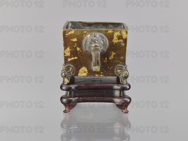 Censer on base, Chinese, Qing dynasty 1644–1911, 18th century. Bronze censer with lion fo iconography and handles; ritual/ceremonial vessel. Wooden carved quadrangular base. Dimensions: censer height 2.2 cm, width 12.3 cm, length 9.7 cm; base height 1.8 cm, width 12.2 cm, length 9.6 cm., A square metal vessel with a worn gold and brown surface sits on a dark reddish-brown wooden stand; the vessel shows patches where the gold finish is flaking, and a metallic lion head ornament with a short pendant is affixed to the front, flanked by decorative circular elements on either side.