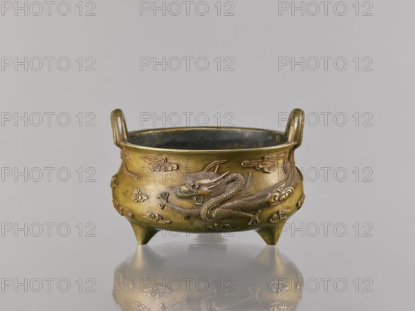 Li-type incense burner censer, Chinese, 18th century copy after a 15th-century original, Qing dynasty 1644–1911. Bronze vessel, height 11.5 cm, width 17.6 cm. Decoration: two dragons among clouds; motifs include dragons and clouds. Ceremonial/ritual ding-style vessel; backdated signatures., A small bronze tripod vessel with a round bowl and two upright loop handles, resting on three short conical legs, shown against a plain light gray background; the vessel surface is a warm golden-bronze with darker brown and greenish patina in places, and it is decorated with raised dragon motifs and stylized cloud-like forms in the same metal finish; the interior appears darker with a bluish-green tinge, and the vessel casts a soft reflection on the glossy surface beneath it.