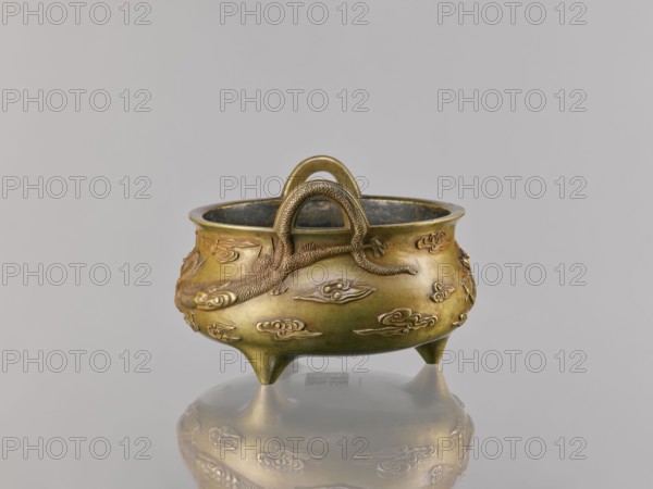 Li-type incense burner, ding vessel form, Chinese art; dragons and clouds iconography; ceremonial/ritual vessel; Qing dynasty 1644–1911, 18th century copy after a 15th-century original; backdated signatures; Decoration: two dragons among clouds; vessel censer; height 11.5 cm, width 17.6 cm., A small rounded metal vessel with a warm golden-bronze color sits on a reflective surface, showing a faint mirrored image beneath; the vessel has a wide rim and a slightly darkened interior, and rests on three short pointed legs. A textured handle arches over the top, and a raised snake or dragon-like figure wraps around the body, rendered in the same golden-bronze tone with fine scale details. The exterior surface is decorated with several low-relief cloud-shaped motifs in matching bronze, and the overall background is a smooth, pale gray.