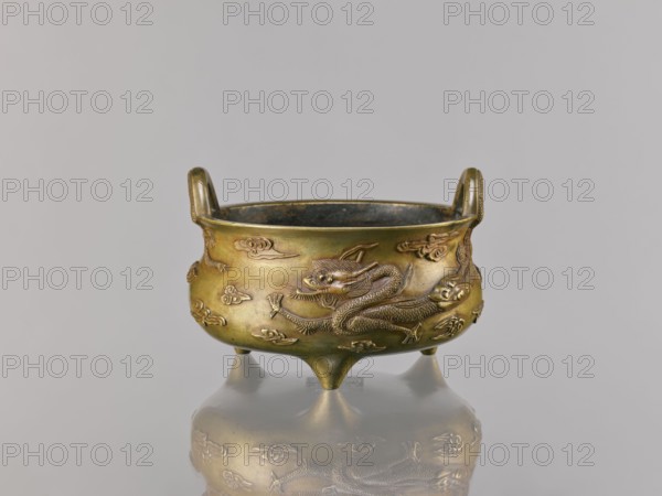 Li-type incense burner, ; ding vessel; Chinese; Qing 1644–1911, 18th century copy after a 15th-century original; ceremonial/ritual censer vessel; material likely bronze; decoration: two dragons among clouds; dragons iconography; clouds motif; backdated signatures; height 11.5 cm, width 17.6 cm., An antique metal vessel with a rounded body and two upright handles sits on three short conical legs, showing a raised dragon motif and decorative cloud-like reliefs around its exterior; the vessel appears in shades of gold, brass, and muted bronze with darker interior tones, and it is reflected on a smooth grayish surface beneath it against a light gray background.