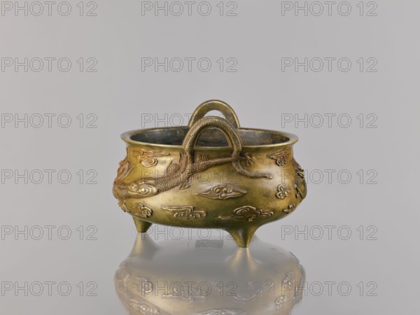 Li-type incense burner censer, Ding-style vessel, Chinese, 18th century copy after a 15th-century original, Qing dynasty 1644–1911. Bronze censer, height 11.5 cm, width 17.6 cm. Decoration: two dragons among clouds; backdated signatures; ceremonial/ritual function., A small metal vessel with a rounded body and three short conical legs sits on a reflective surface, showing a mirrored image beneath. The vessel is primarily brass-colored with areas of darker patina and greenish tones inside the rim. Raised decorative elements in the same metallic hues cover the exterior, including a textured rope-like handle arching over the top and a sculpted long animal motif wrapping around the body. The background is a smooth light gray, and the reflection on the surface echoes the vessel's brass and darker greenish colors.