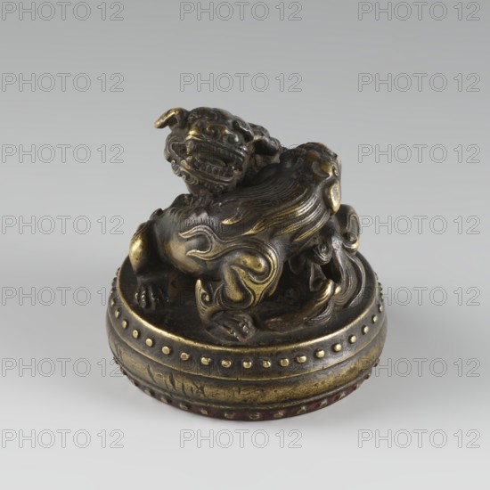 Seal stamp, fo iconography, buttons, Qing style, zoomorphic motifs, Chinese culture, Chinese art, lions iconography, painting implements, Qing 1644–1911, 19th century, China, seal stamp, height 6.2 cm, diameter 5.7 cm, A small sculptural object shows a stylized crouching creature with a broad, open-mouthed face, curled mane, and curled tail, positioned atop a rounded base with a decorative rim. The creature and base are metallic with a mix of dark bronze and brighter gold tones, showing areas of patina and wear. The base has a band of raised rounded studs around its upper edge. The background is a smooth, light gray gradient.