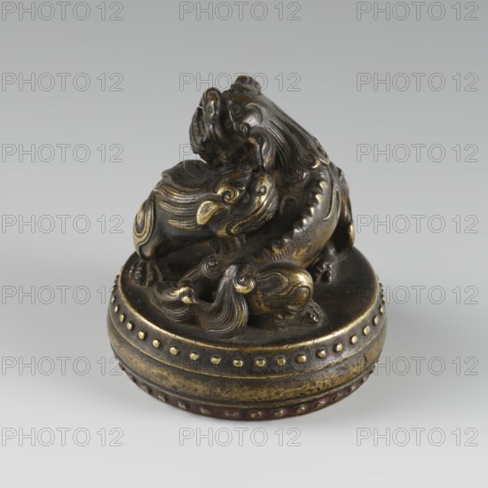 Seal stamp, fo iconography, buttons, Qing style, zoomorphic motifs, Chinese culture, Chinese art, lions iconography, painting implements, Qing 1644–1911, 19th century, China, seal stamp, height 6.2 cm, diameter 5.7 cm, A small sculpted object sits on a round base, showing a curled mythical creature with flowing, stylized fur and a sinuous body. The piece displays a metallic surface with a dark brown patina and highlights of golden tones on raised areas and decorative swirls. The circular base has a banded design with a row of raised dots and worn areas revealing darker brown and muted reddish accents. The background is a smooth, light gray that contrasts with the darker brown and gold of the object.