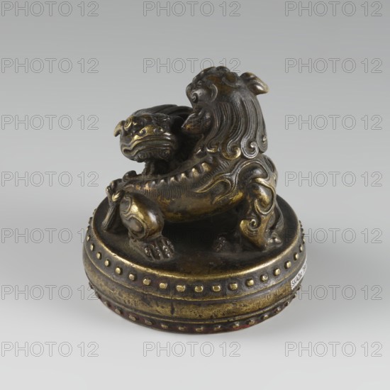 Seal stamp, ; fo iconography; buttons; Qing style; zoomorphic motifs; Chinese culture; lions iconography; painting implements; Qing 1644–1911, 19th century, China; seal stamp; height 6.2 cm, diameter 5.7 cm, A small sculpted object depicting a stylized animal with a curled mane and muscular limbs sits atop a rounded base; the figure has a broad snout, curled tail, and decorative carved swirls along its body, with a companion head positioned near its side. The piece displays a metallic surface with a patina showing shades of dark bronze and areas of golden highlights, along with touches of darker brown and subtle reddish tones on the lower rim, all set against a neutral light gray background.