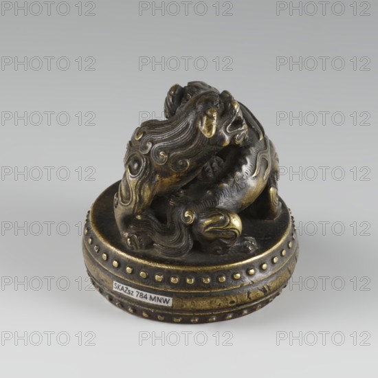 Seal stamp, fo iconography, buttons, Qing style, zoomorphic motifs, Chinese culture, Chinese art, lions iconography, painting implements, Qing 1644–1911, 19th century, China, seal stamp, height 6.2 cm, diameter 5.7 cm, A small sculptural object sits on a round base, showing a stylized animal-like figure with flowing, carved mane and curled decorative patterns; the piece is metallic with visible gold and dark bronze tones, and a patinated surface showing areas of shine and darker wear.