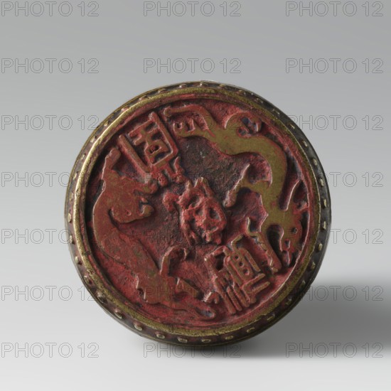 Seal stamp, fo iconography, buttons, Qing style, zoomorphic motifs, Chinese, lions iconography, painting implements, Qing 1644–1911, 19th century, China, seal stamp, height 6.2 cm, diameter 5.7 cm, A round object with a raised rim and a textured circular face, showing a worn sculpted central relief with organic, curving shapes and carved figures; the surface displays predominant red and dark red tones with areas of gold and brass-colored highlights on the rim and parts of the relief, and a few darker brown and blackened spots from age or patina, all set against a neutral light gray background.