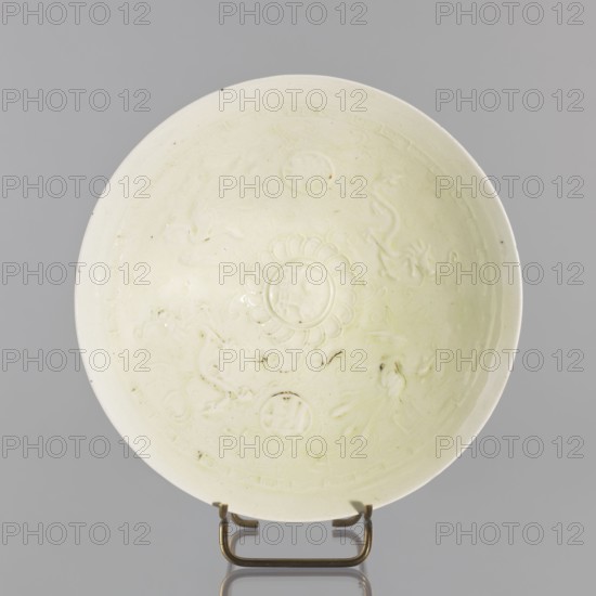 Rice bowl, Yuan dynasty 1271–1368, late 13th century, China; ceramic vessel decorated with rosettes, dragons and phoenixes fenghuang; height 6.5 cm, diameter 19.8 cm; used for rice., A shallow round dish with a pale creamy off-white glaze displayed upright on a small metal stand, showing a central raised floral medallion surrounded by shallow embossed decorative motifs and curved ornamental designs, with subtle variations in glazing tone and a few small darker specks and spots on the surface, set against a neutral gray background; the metal stand is a brass-like color.