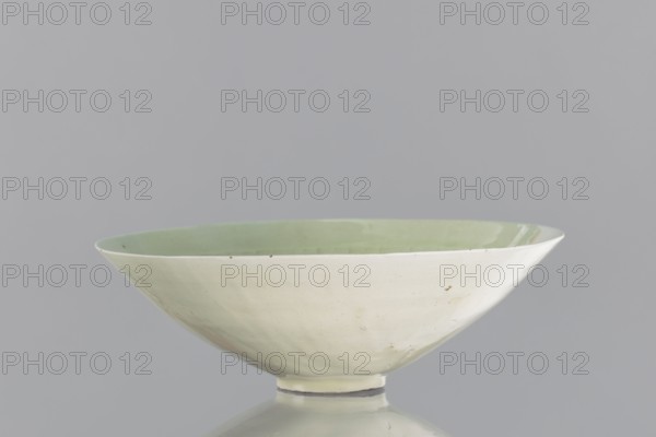 Rice bowl, purchased provenance; Chinese art; culture: Chinese; Yuan dynasty 1271–1368; ceramics; bowl for rice; decoration: rosettes, dragons, phoenixes fenghuang; late 13th century; China; vessel; height 6.5 cm; diameter 19.8 cm, A shallow, wide ceramic bowl with a pale green glossy interior and a creamy off-white exterior, sitting on a small circular foot; the bowl shows subtle surface imperfections and tiny dark specks, and it rests on a reflective surface against a neutral light gray background.
