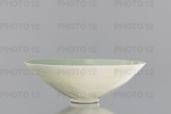 Rice bowl, Yuan dynasty 1271–1368, late 13th century, China; ceramics vessel for rice; decorated with rosettes, dragons, phoenixes fenghuang; height 6.5 cm, diameter 19.8 cm., A shallow ceramic bowl with a pale green interior and an off-white exterior is shown against a gray background, sitting on a reflective surface that mirrors the bowl's shape; the bowl has a slightly irregular rim and a small foot at the base, and the visible colors include pale green, off-white, gray, and a hint of darker specks on the glaze.