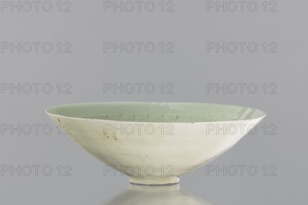 Rice bowl, Yuan dynasty 1271–1368, late 13th century, China; stoneware ceramic vessel decorated with rosettes, dragons and phoenixes fenghuang; function: rice bowl; height 6.5 cm, diameter 19.8 cm., A shallow, wide bowl with a pale green interior and an off-white exterior sits centered against a neutral gray background; the bowl has a smooth, slightly glossy surface with a few small blemishes and subtle variations in tone, a narrow foot at its base, and a gentle curved profile forming the rim.