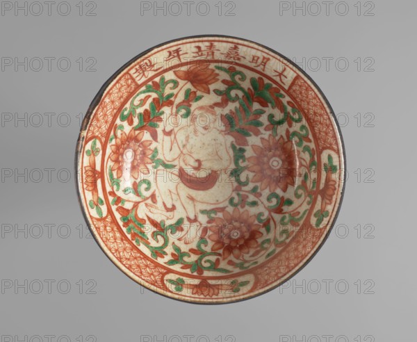 Bowl, Chinese, Ming dynasty, Jiajing 1522–1566, colored porcelain, decorated with phoenixes fenghuang, dragons long, lotus tendril ornament, and boys. Decoration: inside — figure of a naked boy with a lotus flower among lotus tendrils; outside — feng bird and long dragon among lotus tendrils; framed at top and bottom with a meandering border. Fragment is thick, stoneware-like. Date: ca. 1522–1566. Place of origin: China. Vessel; height 7 cm, diameter 18.3 cm., A shallow round bowl viewed from above with a central figure seated among scrolling floral vines; the decoration features a palette of red-orange, green, and cream with darker brown outlining; the rim has a patterned band and panels of geometric and floral motifs in red-orange and green; several large stylized flowers and leafy tendrils fill the interior surrounding the figure; the background of the bowl is a crackled cream color and the outer edge has a thin darker brown rim; overall colors visible are red-orange, green, cream, ...