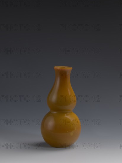 Vase, Chinese, Qing dynasty 1644–1911, 18th century. Double-gourd form, yellow opaque glass, cast and cut, stained-glass appearance; height 16.5 cm, diameter 8.0 cm, weight 0.41 kg., A glossy gourd-shaped vase in a warm amber color sits centered against a smooth gradient background that transitions from dark gray at the top to lighter gray near the bottom, with a soft shadow beneath the vase and subtle highlights and reflections on its curved surfaces.