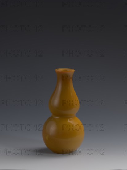 Vase;, Chinese, Qing dynasty 1644–1911, 18th century. Double-gourd form vessel made of yellow glass, cast and cut, opaque; stained glass effect. Height 16.5 cm; diameter 8.0 cm; weight 0.41 kg., double gourds, gourds, vases., A glossy gourd-shaped vase in a warm yellow-orange color sits centered against a smooth gradient background that transitions from dark gray at the top to lighter gray near the bottom; the vase has a rounded lower bulb, a narrower midsection, and a flared small neck with visible highlights and soft reflections, casting a subtle shadow on the light gray surface beneath it.