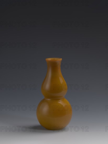 Vase, Chinese, Qing dynasty 1644–1911, 18th century. Double gourd-shaped vessel made of yellow glass, cast and cut, opaque; stained glass effect. Height 16.5 cm; diameter 8.0 cm; weight 0.41 kg., A smooth, glossy gourd-shaped vase in a warm amber color sits centered on a surface, showing reflective highlights and subtle shadows; the background transitions from a dark gray at the top to a lighter gray near the bottom, and the surface beneath the vase is a pale gray with a soft shadow on one side.