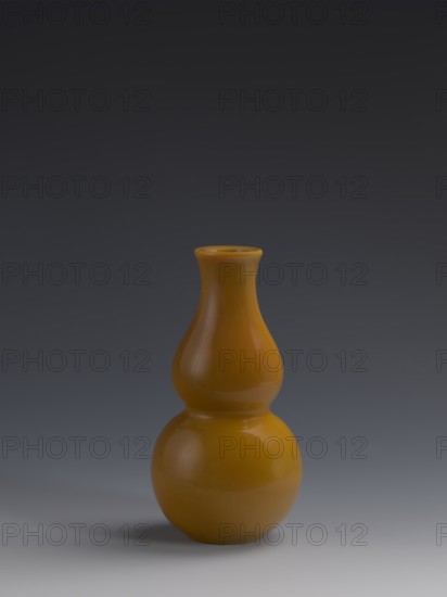 Vase, Chinese, Qing dynasty 1644–1911, 18th century. Yellow glass, cast and cut, opaque; double gourd form. Vessel with height 16.5 cm, diameter 8 cm, weight 0.40 kg., A glossy gourd-shaped vase with a bulbous lower section and a narrower upper neck sits centered against a smooth gradient background; the vase is a warm mustard yellow color with highlights and soft reflections, and the background transitions from dark charcoal at the top through slate gray to a lighter gray near the bottom, where a soft shadow under the vase appears in muted gray tones.